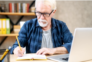 man in flannel shirt in front of laptop taking notes in book