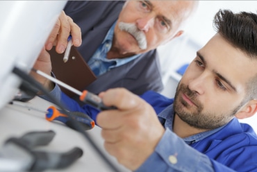man with white mustache trains younger man with tools in foreground