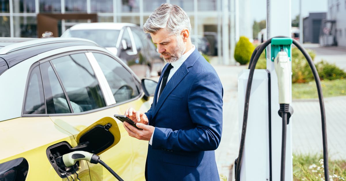 Mature Caucasian businessman using a smartphone while waiting for his electric vehicle to charge at a workplace EV charging station outdoors