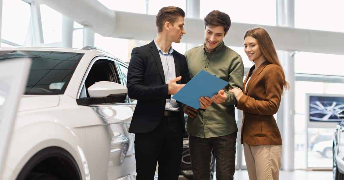 A joyful young couple discussing with a salesman at an automobile dealership, signifying the excitement and considerations when investing in a second EV