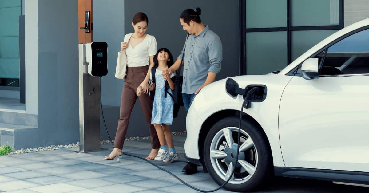 Young, progressive parents and their daughter stepping out of their home, with an electric car charging in the driveway, showcasing the real-life application of installing an electric vehicle charging station at home