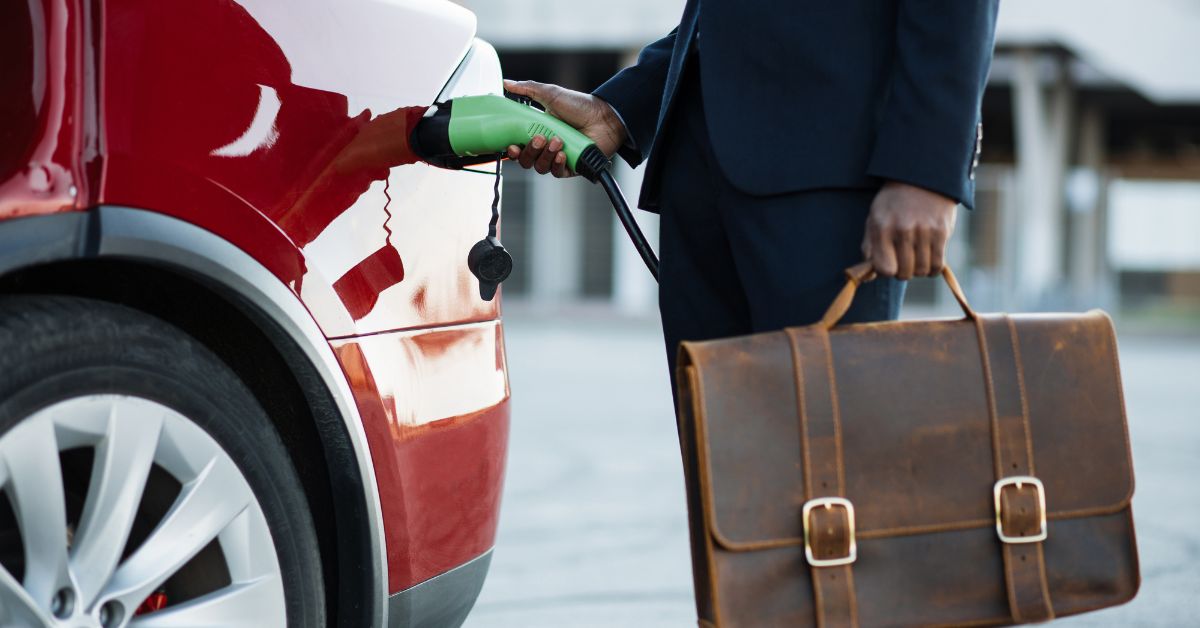 A gentleman in a formal suit connects a charging cable to an electric car, holding a leather suitcase, symbolizing the economic and environmental advantages of adopting a fleet of electric vehicles