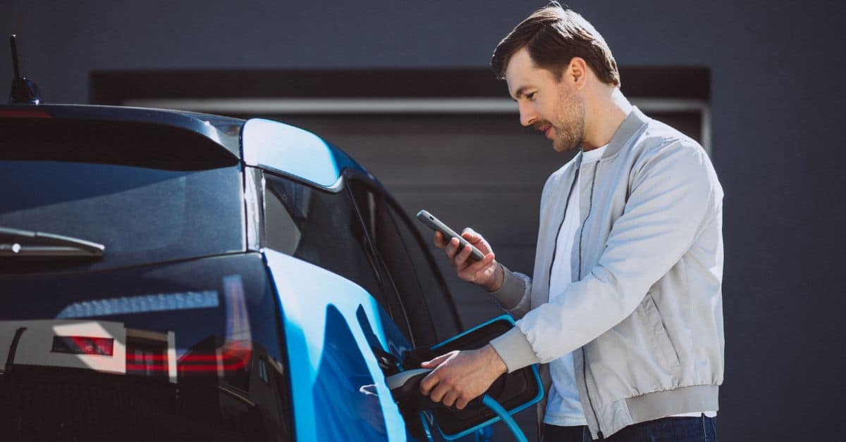 A young man charging his electric car at home and checking his mobile phone, representing the variables that impact home EV charging speed