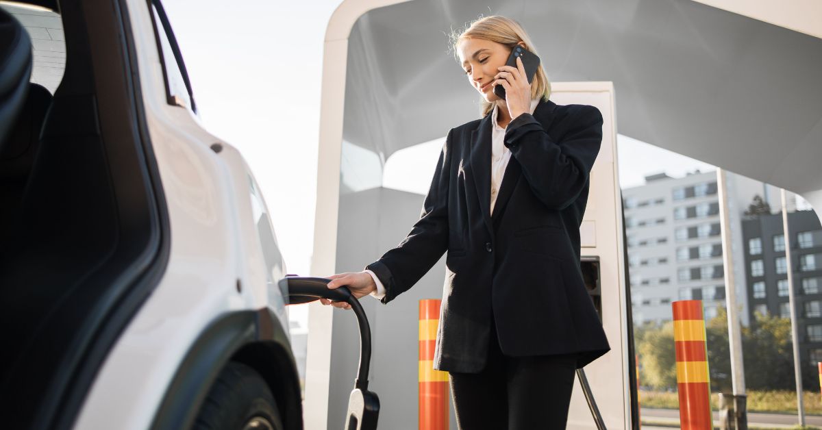 A businesswoman charging her electric vehicle outdoors, talking on the phone. Illustrating practical electric vehicle fleet maintenance strategies in a corporate setting.