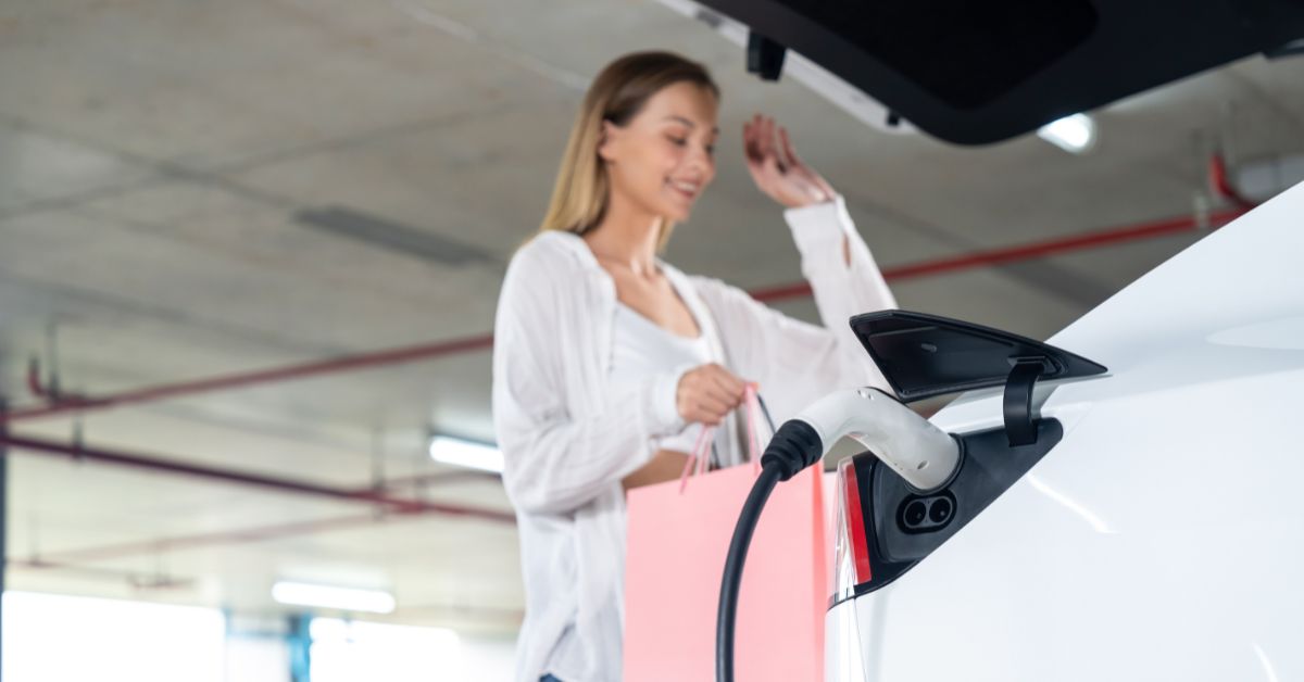 A young woman beside her charging EV at a shopping center, underlining the role of commercial EV charging stations, relevant for building owners